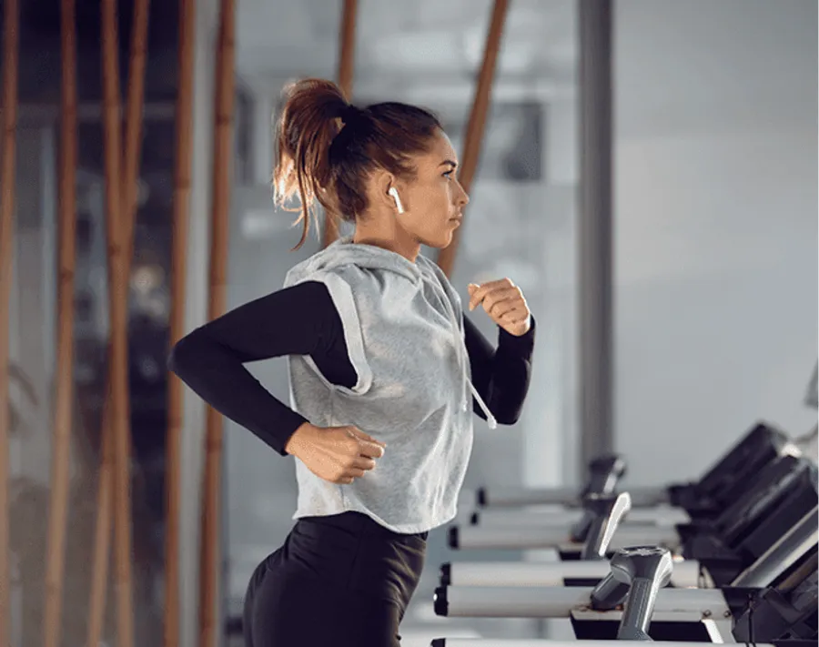Woman in workout clothes running on a treadmill in a gym wearing wireless earbuds.
